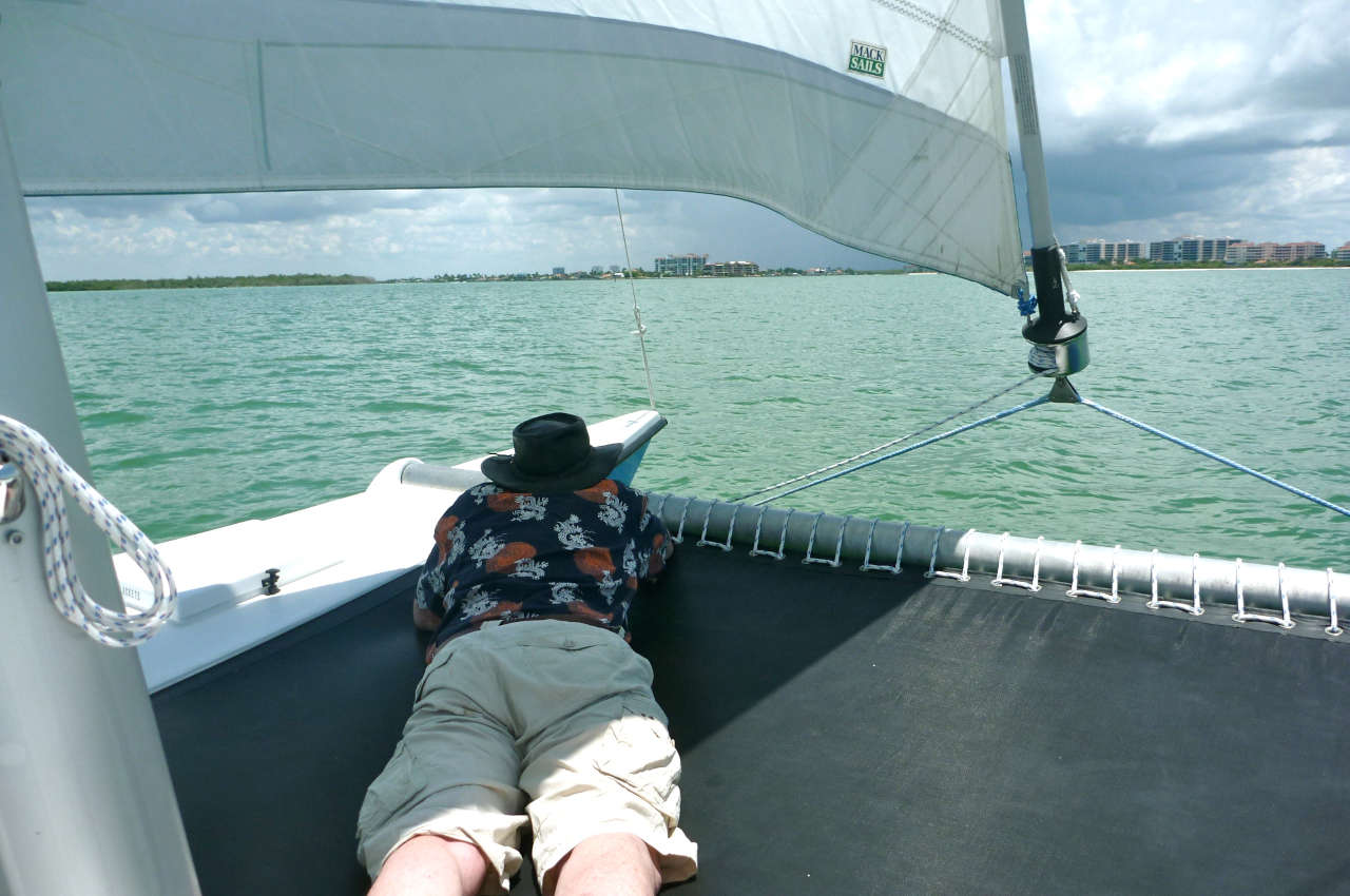 A man lying on the bow trampoline of a catamaran, peering out into the water