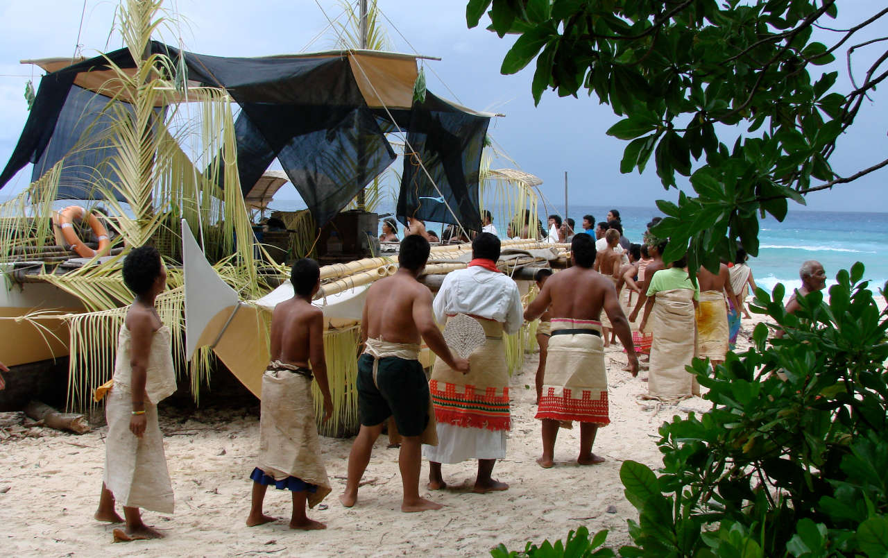 A large group of people in sarongs walking around a beached double canoe, which is decorated in palm leaves